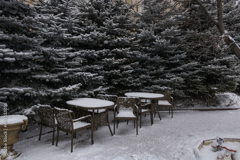 Snow-covered tables and chairs of the outdoor cafe next to the fir trees. An empty cafe. No people. Winter in the southern city of Russia.