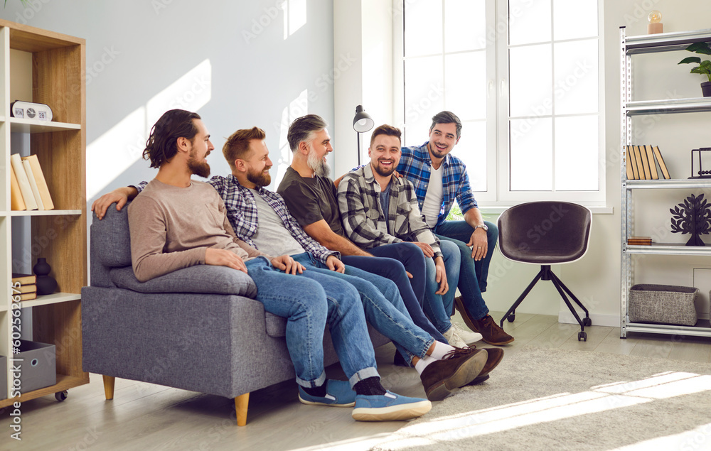 Fototapeta premium Portrait of a male bearded friends sitting on sofa, smiling and having fun at home in the living room. Photo of group of brutal friendly men in casual clothes talking indoors on the party.