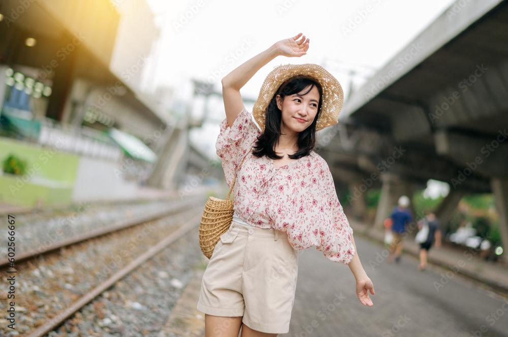 young asian woman traveler with weaving basket happy smiling looking to a camera beside train railway. Journey trip lifestyle, world travel explorer or Asia summer tourism concept.