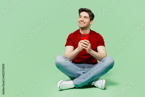 Full body smiling happy young man he wears red t-shirt casual clothes hold in hand use mobile cell phone look aside isolated on plain pastel light green background studio portrait. Lifestyle concept.