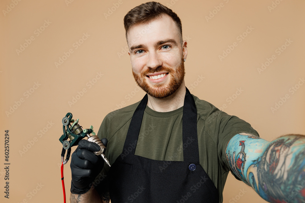 Φωτογραφία Close up tattooer master man wears green t-shirt apron do selfie shot pov mobile cell phone hold machine black ink in jar, equipment for making tattoo art on body isolated on plain beige background