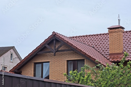 brown brick attic of a private house with windows under a tiled roof with a chimney behind a fence on the street against a gray sky