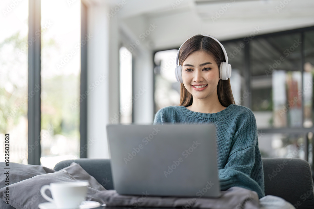 Young woman in modern headphones sit on couch in living room work on laptop Young woman in modern headphones sit on couch in living room work on laptop