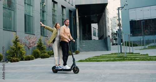 Happy young guy and girl ride on electric scooters near business center at street outdoors, after work. Lifestile concept.