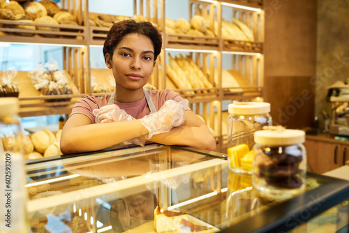 Young pretty female clerk standing by glass display with pastry in bakery shop and looking at camera against shelves with fresh bread