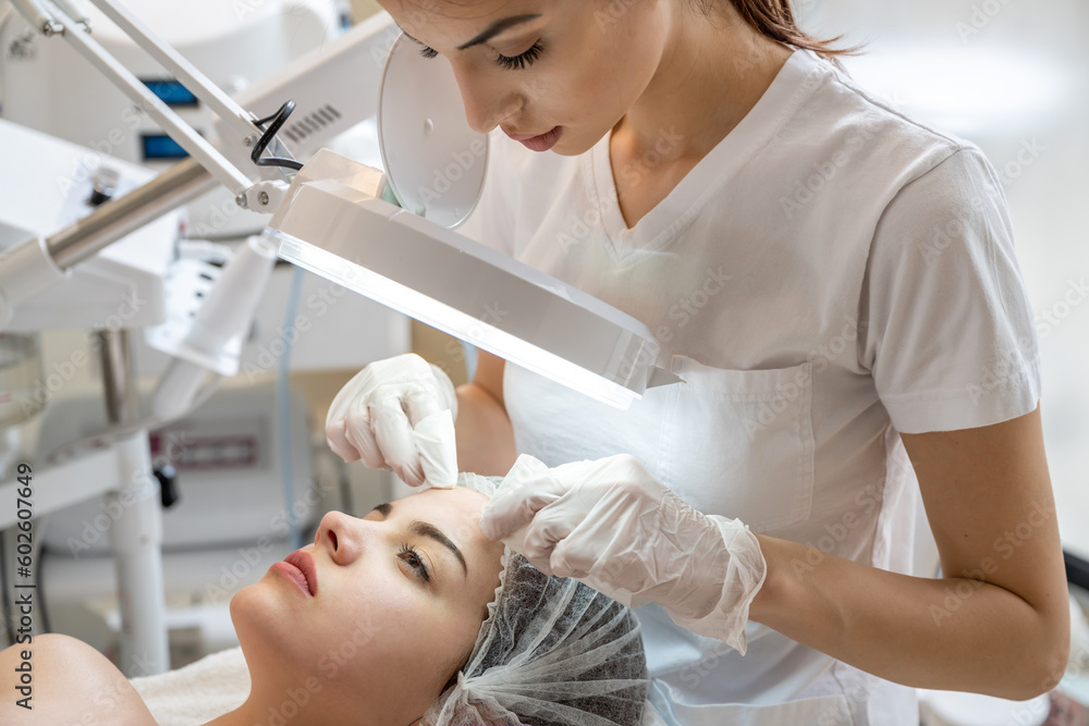 Beautician squeezing acne and pimples from young woman's nose using lamp and magnifying glass in beauty spa clinic.