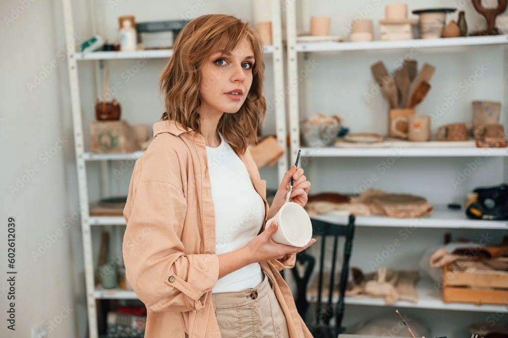 Beautiful young woman is standing and painting a ceramic pot in the workshop