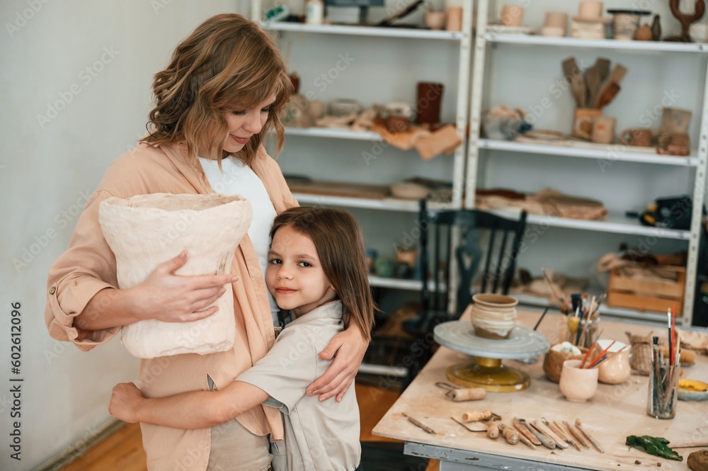 Woman is standing with daughter and holding big unfinished ceramic pot