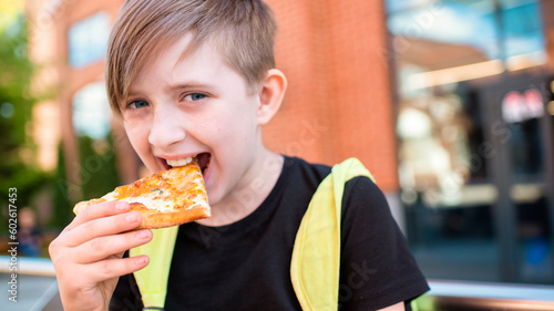  the boy holds a slice of pizza in his hand  is going to eat. the child ordered pizza  sits on a bench with pizza in a box on a sunny day in the backyard of the school