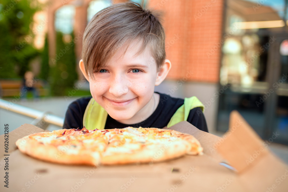 the boy holds  pizza, is going to eat. the child ordered pizza, sits on a bench with pizza in a box on a sunny day in the backyard of the school