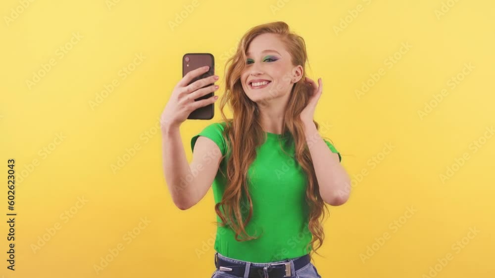 Attractive red-haired woman takes a selfie on the front camera of her smartphone. A young woman in a green T-shirt stands on an orange background with a phone in her hand. Attractive red-haired woman takes a selfie on the front camera of her smartphone. A young woman in a green T-shirt stands on an orange background with a phone in her hand.