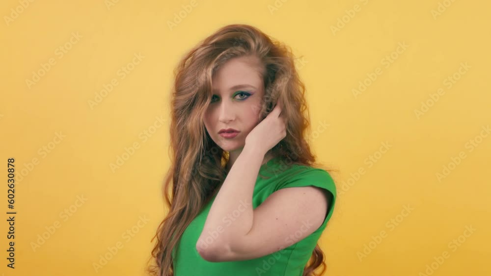 Beautiful woman with red hair posing for the camera. A young long-haired woman in a green t-shirt stands against an orange wall and straightens her hair while looking at the camera. Beautiful woman with red hair posing for the camera. A young long-haired woman in a green t-shirt stands against an orange wall and straightens her hair while looking at the camera.