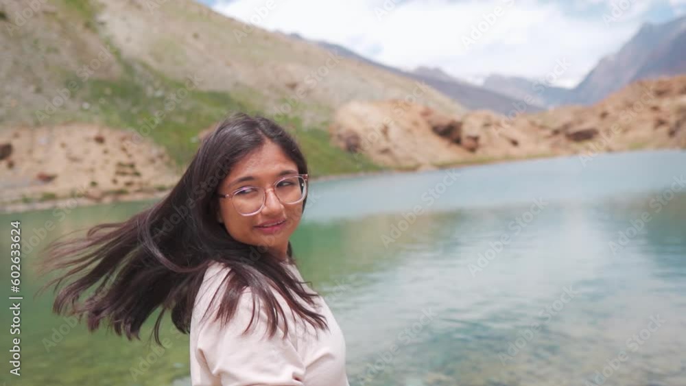 Portrait of beautiful Indian girl looking back facing camera, hair waving in cold wind at Deepak Tal Lake in Lahaul, India. Girl standing against mountains and glacier water lake. Travel lifestyle