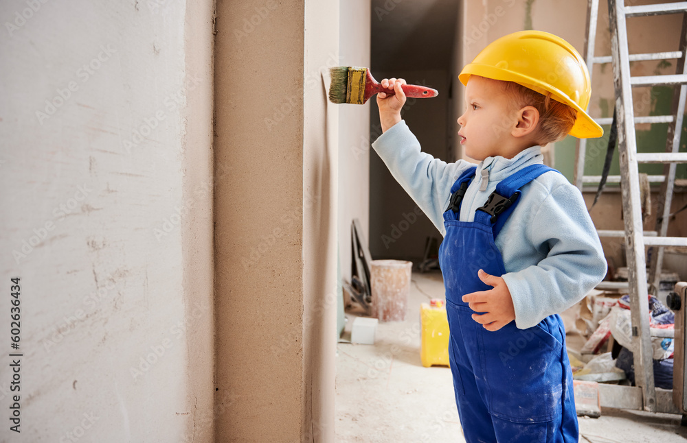 Little kid construction worker painting wall with brush in apartment under renovation. Child wearing safety helmet and work overalls while working on home renovation. - obrazy, fototapety, plakaty 
