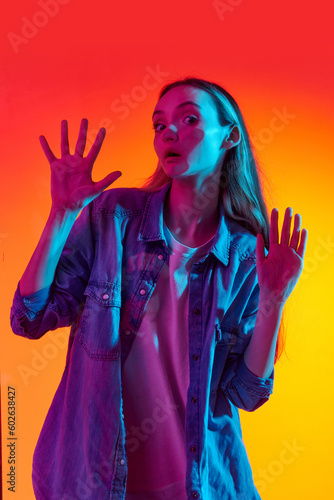 Portrait of one young, adorable girl, teenager looking at camera through glass with shocked face over red studio background in neon light. Funny meme