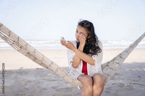 Hispanic woman is using a spoon to do her eyelashes on a hammock at the beach photo