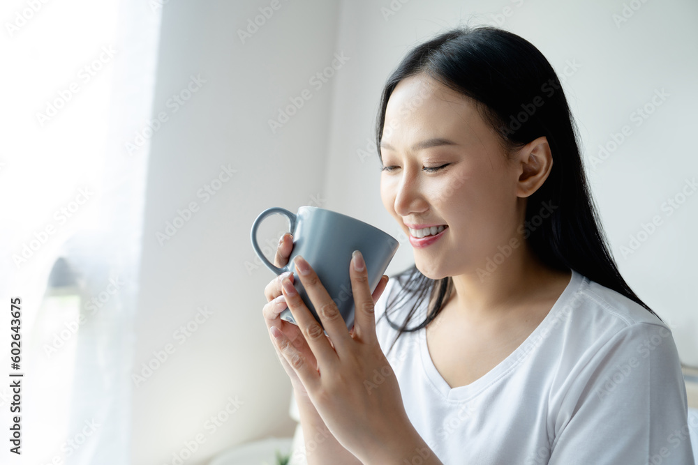 Closeup image of Attractive smiling young woman with long black hair holding coffee mug sitting on bed in morning, she just woke up Closeup image of Attractive smiling young woman with long black hair holding coffee mug sitting on bed in morning, she just woke up