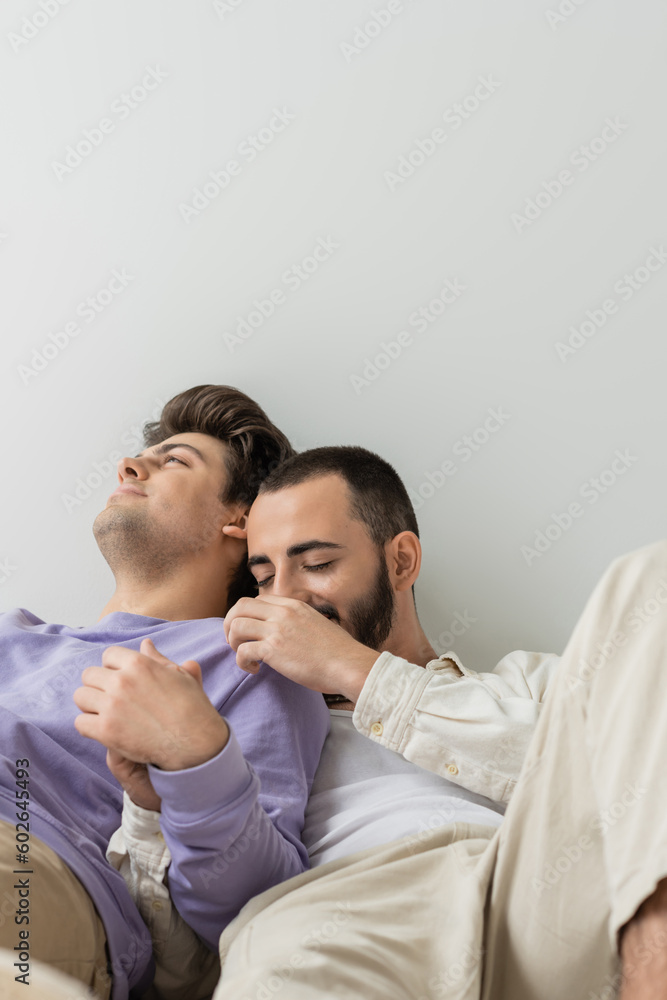Fototapeta premium Young homosexual man touching shoulder and holding hand of brunette boyfriend in casual clothes while sitting together on grey background