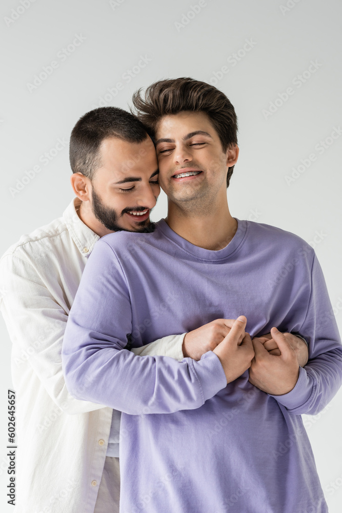 Overjoyed and bearded gay man holding hands and embracing brunette boyfriend with braces and closed eyes while standing isolated on grey Overjoyed and bearded gay man holding hands and embracing brunette boyfriend with braces and closed eyes while standing isolated on grey