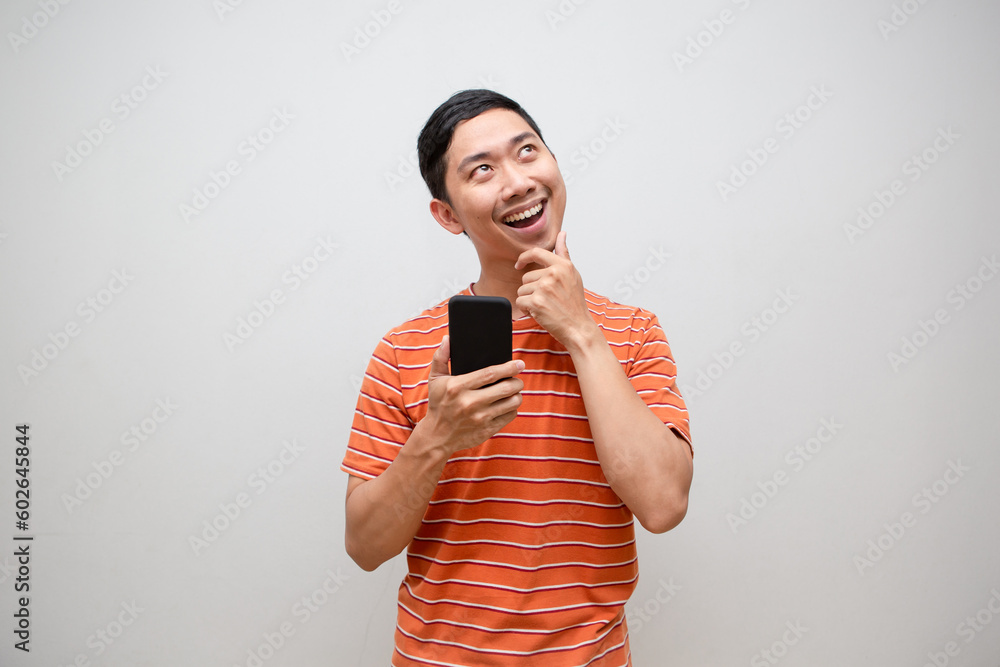 Asian man orange shirt holding smartphone gesture thinking looking up isolated Asian man orange shirt holding smartphone gesture thinking looking up isolated