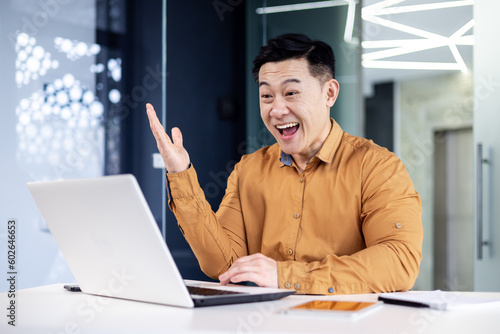 Successful asian man celebrating victory and success received news of good achievement online, businessman looking at laptop screen and holding hand up triumph gesture, worker inside office