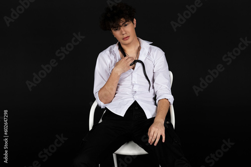 Young handsome man in a white shirt sitting on a chair with a black snake crawling around his neck. Isolated on black background.