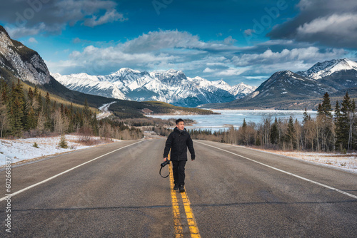 Young asian photographer man walking on the highway with rocky mountains and frozen lake in winter