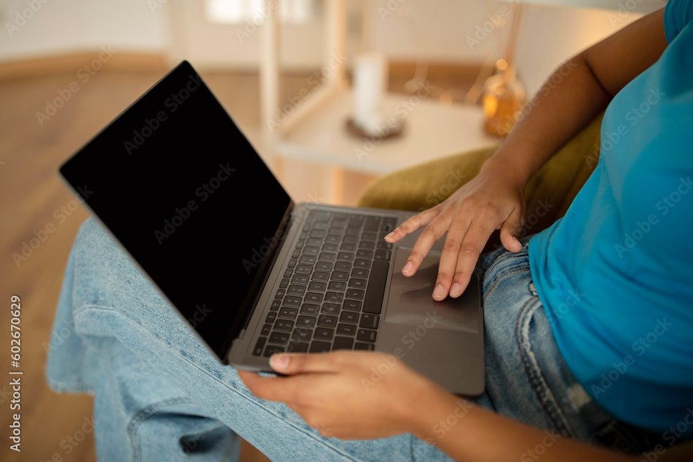 Naklejka premium Millennial curly black lady typing on computer with empty screen in living room interior
