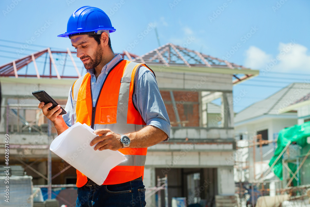 An expert young engineer speaks on the phone. Wears a blue helmet and an orange construction vest. Engineer working on construction site with sunny background An expert young engineer speaks on the phone. Wears a blue helmet and an orange construction vest. Engineer working on construction site with sunny background