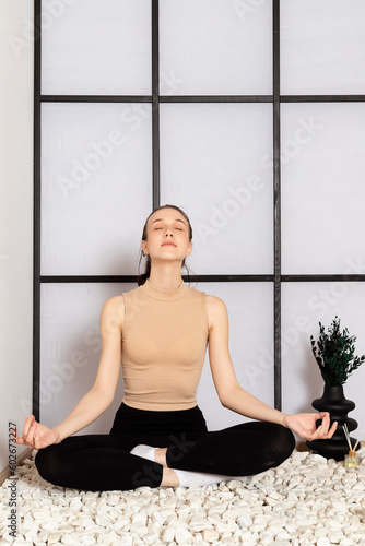 Young Slim brunette Girl practicing yoga, wearing beige sportswear, sitting on the stones, uses incense sticks. Indoor in a Japanese style studio.