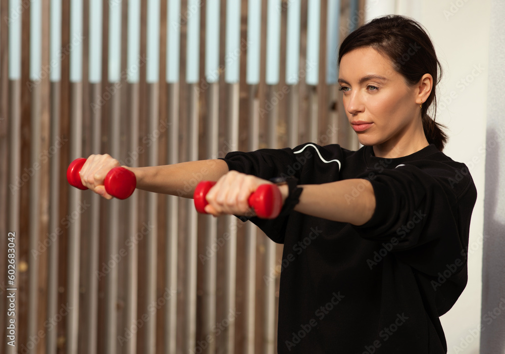 Concentrated millennial european woman athlete in sportswear doing hands exercises with dumbbells