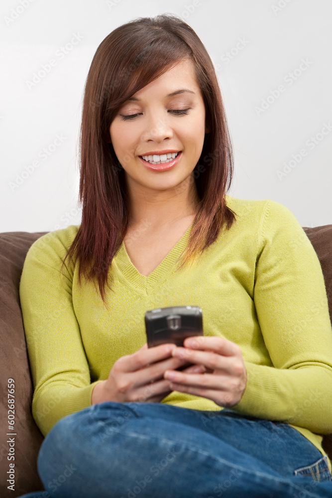 A beautiful young oriental woman with a wonderful toothy smile texting on her smartphone while sitting on a sofa