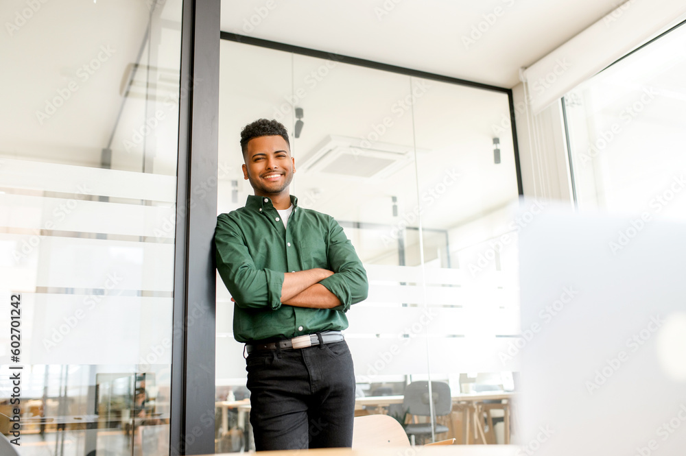 Cheerful indian modern businessman, male entrepreneur standing in contemporary office space in confident pose with folded arms, looks at the camera and smiling, ambitious employee in smart casual wear Cheerful indian modern businessman, male entrepreneur standing in contemporary office space in confident pose with folded arms, looks at the camera and smiling, ambitious employee in smart casual wear