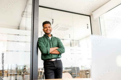 Cheerful indian modern businessman, male entrepreneur standing in contemporary office space in confident pose with folded arms, looks at the camera and smiling, ambitious employee in smart casual wear