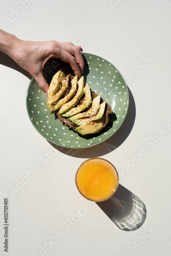 woman's hand holding toast with avocado and sesame next to a glass of orange juice on a white background, detail of a girl eating healthy food for breakfast, bright sunlight photo