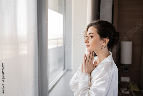 side view of hopeful young woman with engagement ring on finger standing in white silk robe with praying hands and looking through window in hotel suite  special occasion  bride on wedding day