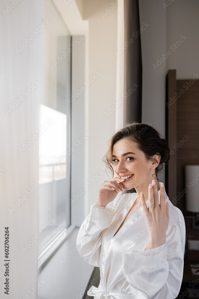 excited young bride with brunette hair in white silk robe showing engagement ring on finger and smiling next to window in hotel suit on wedding day, special occasion, hand near lips excited young bride with brunette hair in white silk robe showing engagement ring on finger and smiling next to window in hotel suit on wedding day, special occasion, hand near lips