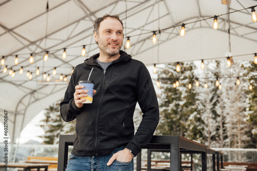 A handsome guy in a black sweater stands with a glass of coffee against the backdrop of mountains and edison lamps A handsome guy in a black sweater stands with a glass of coffee against the backdrop of mountains and edison lamps