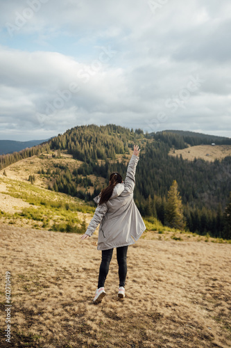 Happy girl stands in the mountains, enjoys the beautiful view and raises her hand up