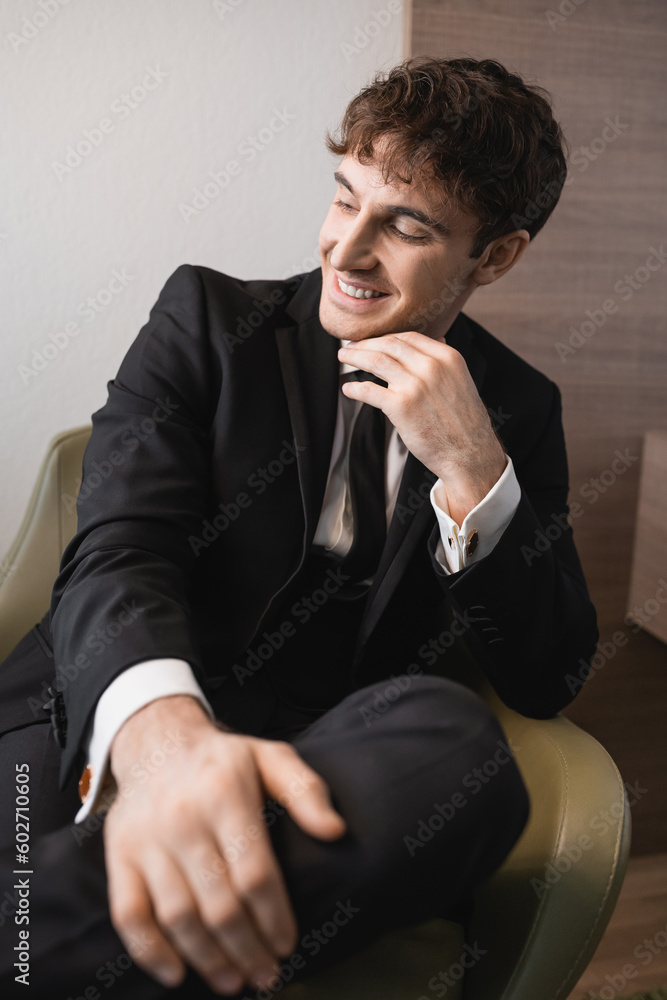 cheerful man in black formal wear with tie sitting on comfortable armchair and looking away on wedding day, resting in modern hotel room, good looking groom cheerful man in black formal wear with tie sitting on comfortable armchair and looking away on wedding day, resting in modern hotel room, good looking groom