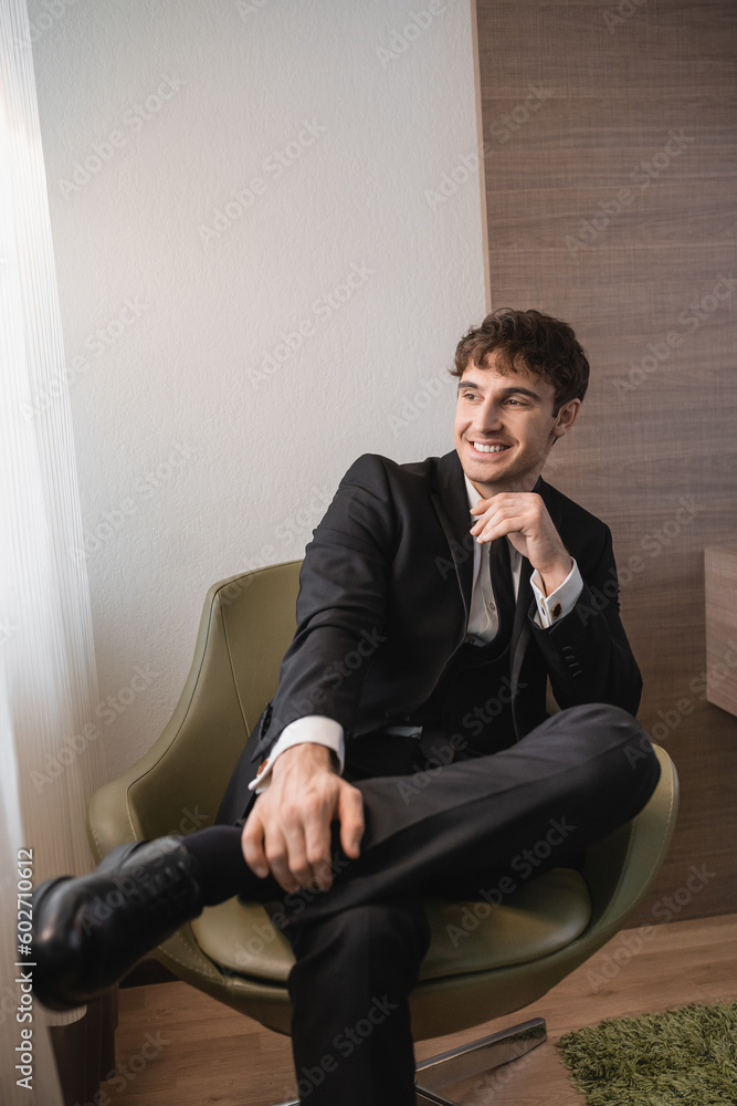 happy man in black formal wear with tie and classic shoes sitting on comfortable armchair and looking away on wedding day, resting in modern hotel room, good looking groom happy man in black formal wear with tie and classic shoes sitting on comfortable armchair and looking away on wedding day, resting in modern hotel room, good looking groom