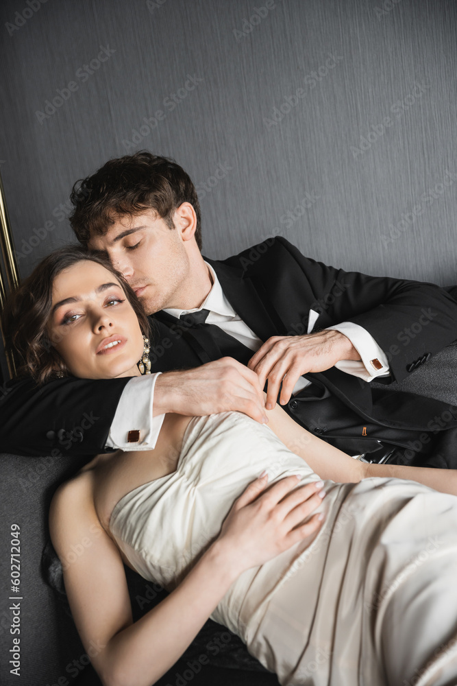 good looking groom in black suit with tie hugging and kissing bride in earrings with pearls and necklace lying in white wedding dress on dark grey couch in hotel room good looking groom in black suit with tie hugging and kissing bride in earrings with pearls and necklace lying in white wedding dress on dark grey couch in hotel room