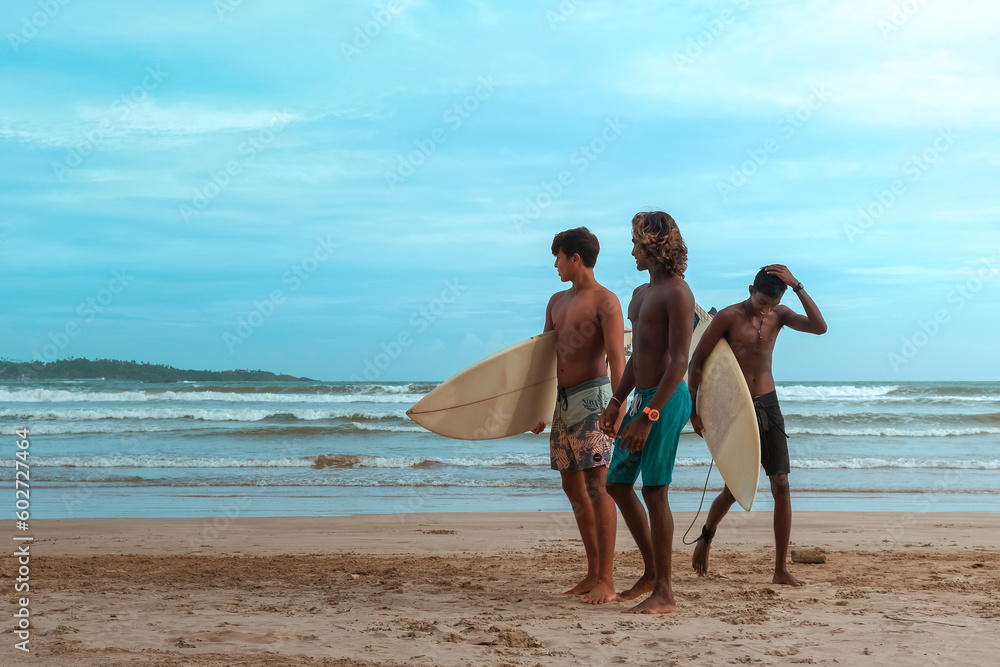 company of young guys Sri Lankans, Indians surfers on the ocean coast with a surfboard, sporty tanned body, smiling and running with a surfboard on the beach on a sunny summer day, ready to surf. company of young guys Sri Lankans, Indians surfers on the ocean coast with a surfboard, sporty tanned body, smiling and running with a surfboard on the beach on a sunny summer day, ready to surf.