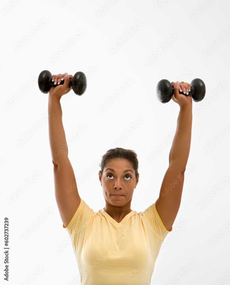 Mid adult multiethnic woman holding dumbbells over head with determined look on her face. Mid adult multiethnic woman holding dumbbells over head with determined look on her face.