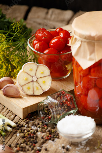 Canned cherry tomatoes in a jar and a bowl, spices and herbs on a wooden background.