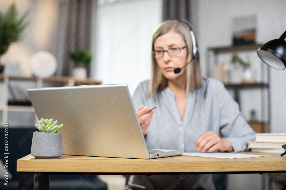 Middle-aged elegant woman using headset with microphone while enjoying home comfort during remote work. Smiling caucasian freelancer in eyeglasses holding online videoconference and making notes. Middle-aged elegant woman using headset with microphone while enjoying home comfort during remote work. Smiling caucasian freelancer in eyeglasses holding online videoconference and making notes.