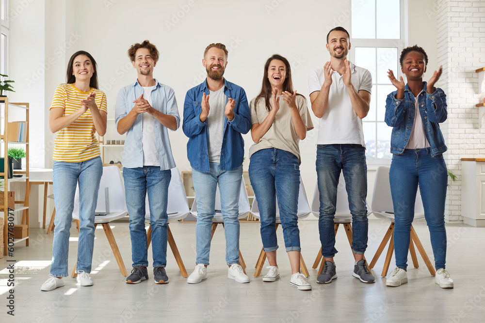 Obraz premium Friendly multiracial people in casual clothes stand in row and applaud looking at you. Group of friends in jeans and T-shirts joyfully clap their hands while standing in row in room. Team concept.