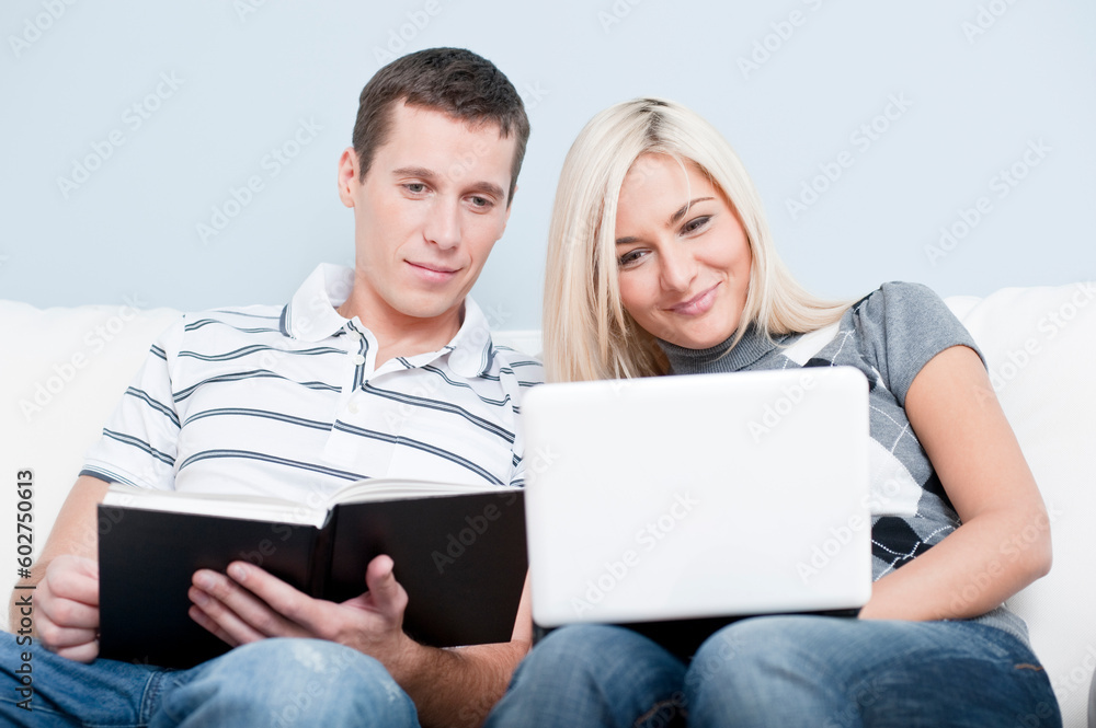 Man is reading, and woman is using a laptop, as they sit side by side on a white couch. Horizontal format.
