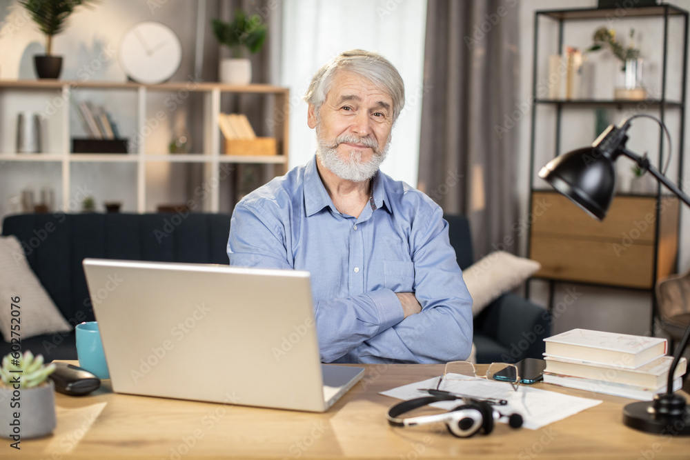 Portrait of senior caucasian man posing with his arms crossed while sitting at office desk with computer and headset. Friendly employee taking break from remote work while staying in cozy home office.