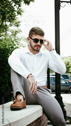 Confident young man wearing sunglasses sitting on restraining wall on college campus. Out of focus cars on a parking lot behind him. photo